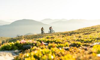Two mountain bikers ride over a green meadow with mountains in the background. The sun is shining and gives the landscape a warm atmosphere.