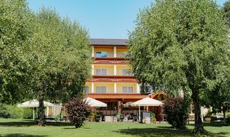 A charming building with three stories, surrounded by green trees. In front of the building are terraces with sun umbrellas.