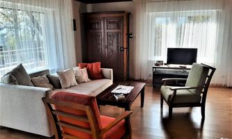 A cozy living space with a white sofa, wooden chairs, and a dark table. Large windows provide plenty of daylight and a friendly atmosphere.