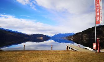 A peaceful landscape with a clear lake and mountains in the background. A dock leads into the water, and in the foreground, a flag is waving with the inscription "BRUNNER".