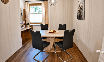 A modern dining area with a wooden table and six black chairs. Natural light streams through the windows, and there is a cozy atmosphere.