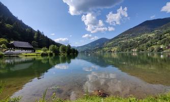 A calm lake surrounded by green mountains and trees. The sky is partly cloudy, and the clouds are reflected in the water.
