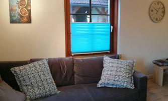 A cozy living area with a gray sofa and colorful cushions. In the background, there is a window with blue blinds and a wall clock.