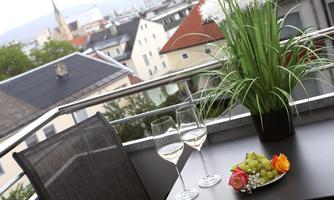 A balcony with a view of the city, equipped with two glasses of wine and a fruit bowl. In the background, green plants and roofs can be seen.
