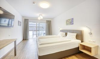 A modern bedroom with two single beds and a light wooden floor. There is a television and access to a balcony through a large glass door.