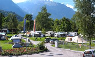 A beautiful campsite with tents and caravans, surrounded by mountains and trees. The scenery is green and inviting, perfect for relaxing in nature.