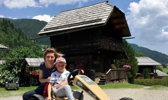 A woman sits with a child in a large go-kart. In the background, traditional wooden houses and a blue sky with white clouds are visible.