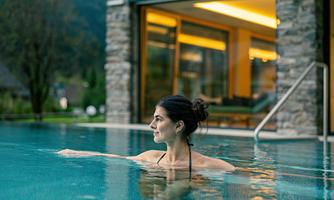 A woman relaxes in the clear water of a pool. In the background, modern architecture and green landscape can be seen.