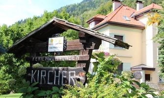 A wooden sign with the inscription "Feriwohnungen Kircher" stands in the foreground. In the background, you can see a modern house surrounded by trees and green landscape.