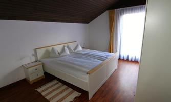 A simple bedroom with a double bed and two pillows. A window with curtains provides light, and a carpet is on the floor.