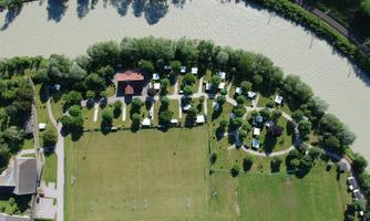 An aerial view of a large campsite by the river. The surroundings are green with trees and meadows, and some RVs are scattered across the site.
