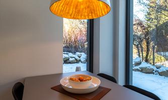 A modern dining room with a large window and a view of a snowy landscape. A bowl of oranges sits on the table, and a stylish lamp illuminates above it.