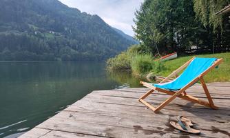 A beautiful, tranquil lake with mountains in the background. A deck chair stands on the wooden walkway and a sandal lies beside it.