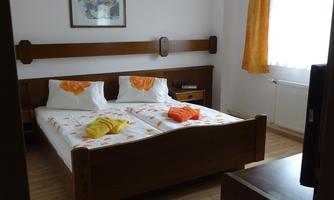 A simple bedroom with a double bed and wooden furniture. The room has a bright atmosphere due to large windows and orange curtains.
