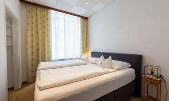 A modern bedroom with two single beds and a large window. The walls are light and the decor is simple and elegant.