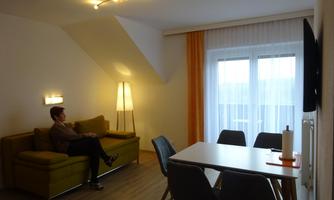 A modern living and dining room with a cozy sofa and a dining table. Large windows provide plenty of daylight.