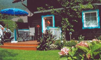A charming wooden house with blue windows and a cozy porch. The green garden features beautiful flowers and a sun umbrella.