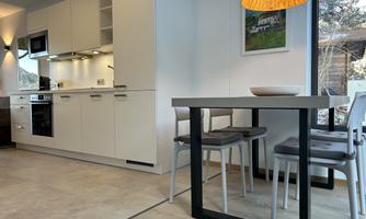 A modern kitchen with white furniture and a dining area. The table has a simple, stylish shape and warm lamp lighting.