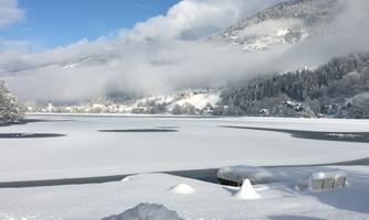 A snowy landscape with a frozen lake and gentle hills in the background. The sky is blue and streaked with some clouds.