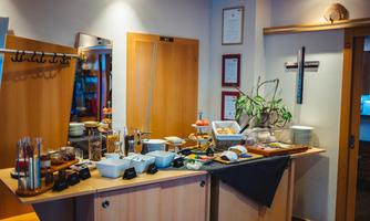 A buffet table with various foods and drinks, including fruits, muesli, and breakfast ingredients. The wall is decorated with pictures and a plant.