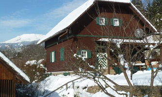 A traditional wooden house in the snow with green windows. Snow-covered mountains can be seen in the background.