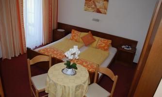 A cozy room with a double bed, pretty bedding, and a small table. The windows are adorned with curtains, and the room feels inviting.