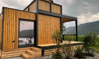 A modern wooden house with large windows and a porch. It is surrounded by green grass and has a view of the mountains.