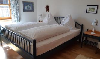 A cozy bedroom with a large double bed and two pillows. The walls are painted in light colors, and there are windows with curtains.