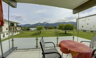 A beautiful balcony with a table and chairs, offering a view of a green meadow and mountains. In the background, a lake is visible and the sky is cloudy.