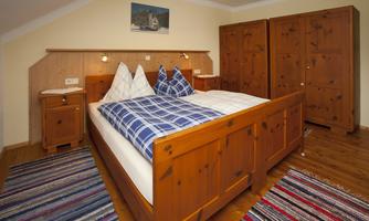 A cozy bedroom with a wooden bed and checked pillows. The room is bright and friendly, with a colorful rug and a wooden cupboard.