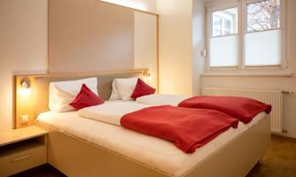 A cozy bedroom with a large bed and red pillows. The walls are light, and there is a window that lets in daylight.