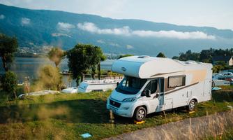A motorhome is parked on a green meadow next to a calm lake. In the background, mountains and a clear sky are visible.