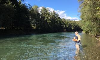A man stands in the clear water of a river, surrounded by trees. The sky is blue with some clouds.