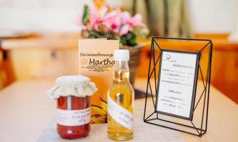 A bottle of oil and a jar of jam are on a table. In the background, there is an information brochure about the vacation apartment "Martha".
