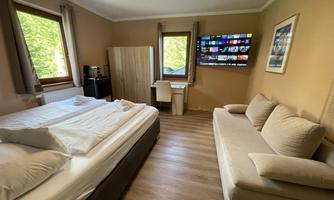 A modern bedroom with a double bed, a sofa, and a large television on the wall. The windows offer a view of greenery and the room is bright and inviting.