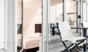 A cozy balcony with a chair and a small table. In the background, a bright room with a bed can be seen.