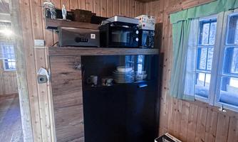 A small wooden room with a black cabinet and a microwave on it. There are windows with green curtains and a rustic wooden floor.