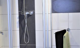 A modern shower with clear, white tiles and a black accent. Towels in various colors hang beside it.