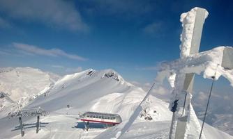A snow-covered mountain landscape with sparkling snow and a chair lift in the foreground. The sky is clear and blue, and the peaks rise majestically.