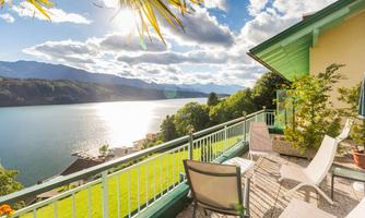 A cozy terrace with a view of a lake and the mountains. The sun is shining and the sky is partially cloudy.