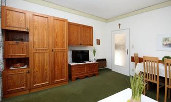 A cozy living room with wooden cabinets, a television, and a dining table. The room has a green carpet and a bright atmosphere.