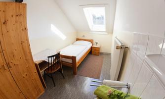 A simple room with a single bed, a desk, and a chair. There is bright light from a skylight and a cozy atmosphere.