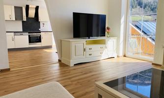 A modern living room with a large television and a beautiful wooden floor. In the background, the kitchen and a window with a view of nature are visible.