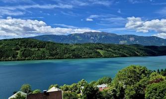 A picturesque landscape with a clear blue lake and green hills. In the background, majestic mountains and a radiant blue sky are visible.
