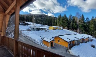 A winter landscape with snow-covered hills and colorful houses. In the background, there are tall trees and a blue sky with clouds.
