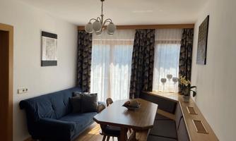 A cozy living room with a blue sofa and a wooden table. Large windows with curtains provide plenty of daylight.