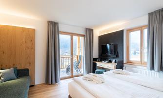 A modern hotel room with two beds and a cozy seating area. Large windows offer a view of a balcony and daylight streams in.