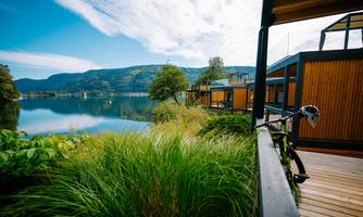 A tranquil lakeside landscape with modern wooden houses by the shore. In the foreground, there is lush grass and a bicycle.