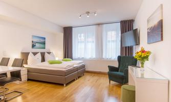 A modern hotel room with a double bed and green pillows. There is a desk, a chair, and windows with daylight.