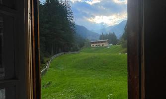 A view from a window of a green meadow and wooded mountains in the background. The sky is partly cloudy and the sun is shining.
